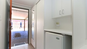 Um corredor estreito com uma porta que dá para área externa, parede de tijolos e chão ladrilhado. Há um frigorífico branco e armários ao longo da parede direita.