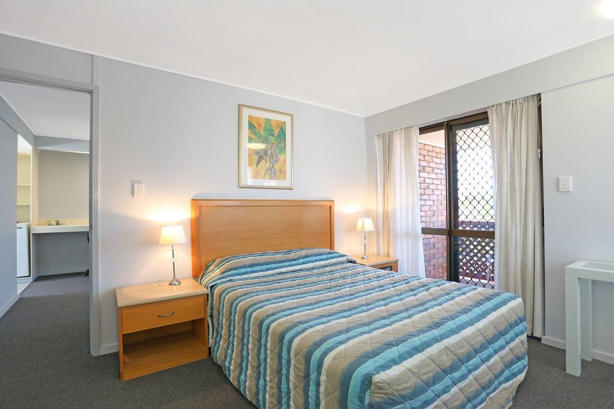 Een slaapkamer met een bed, nachtlampjes, een nachtkastje, een bureau en een raam met gordijnen.