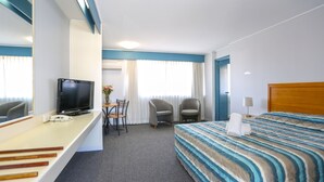 Quarto de hotel com cama, escrivaninha com televisão, duas cadeiras, uma mesinha com um vaso e uma mesinha de cabeceira com abajur.