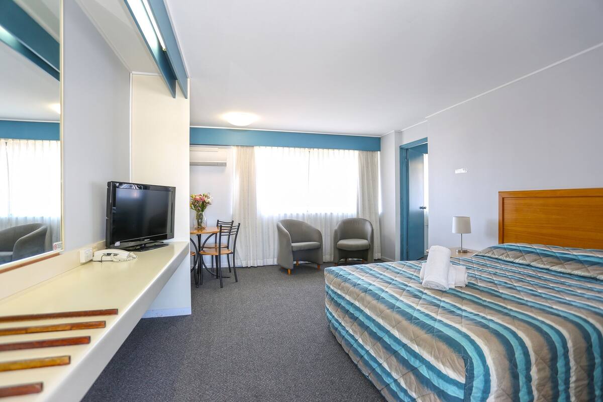 Een hotelkamer met een bed, een bureau met televisie, twee stoelen, een kleine tafel met vaas en een nachtkastje met lamp.