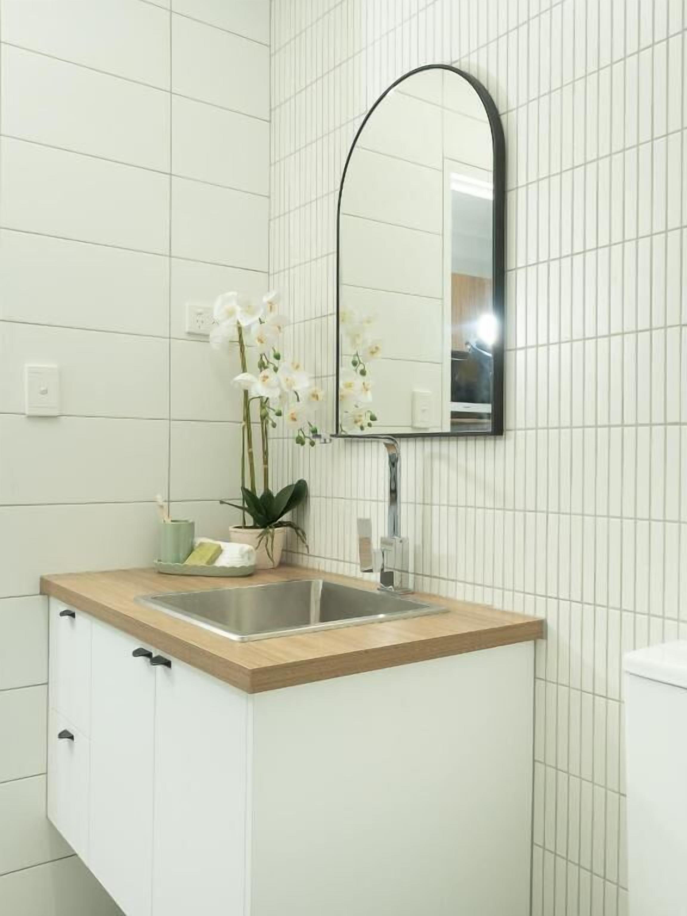 Un baño con pared de azulejos blancos, encimera de madera, lavamanos, espejo y planta en maceta.