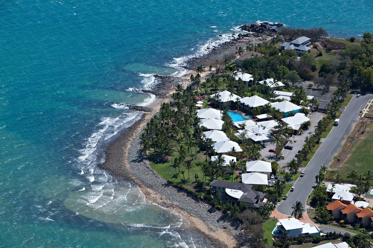 An aerial view of a coastal property with a beach, a swimming pool, and multiple white-roofed buildings.