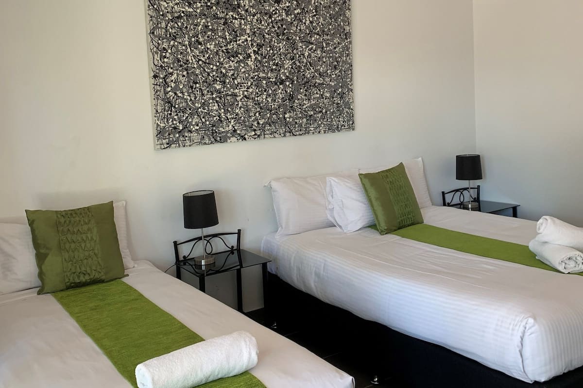 Habitación de hotel con dos camas, cada una con ropa blanca y detalles verdes. Sobre las camas, un cuadro abstracto de gran tamaño.