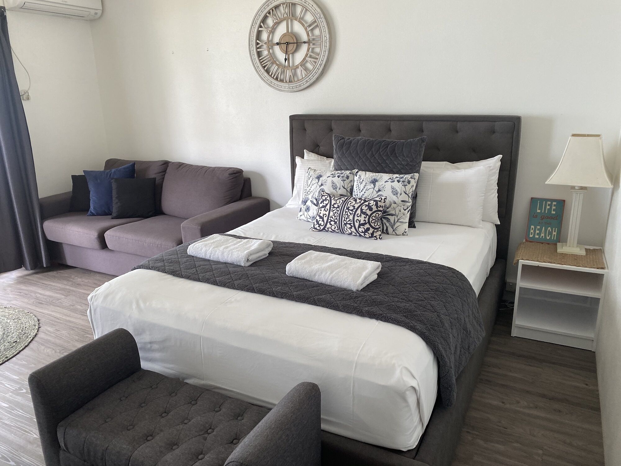 A neatly arranged bedroom with a bed, a sofa, a chair, a bedside table, and a wall clock.