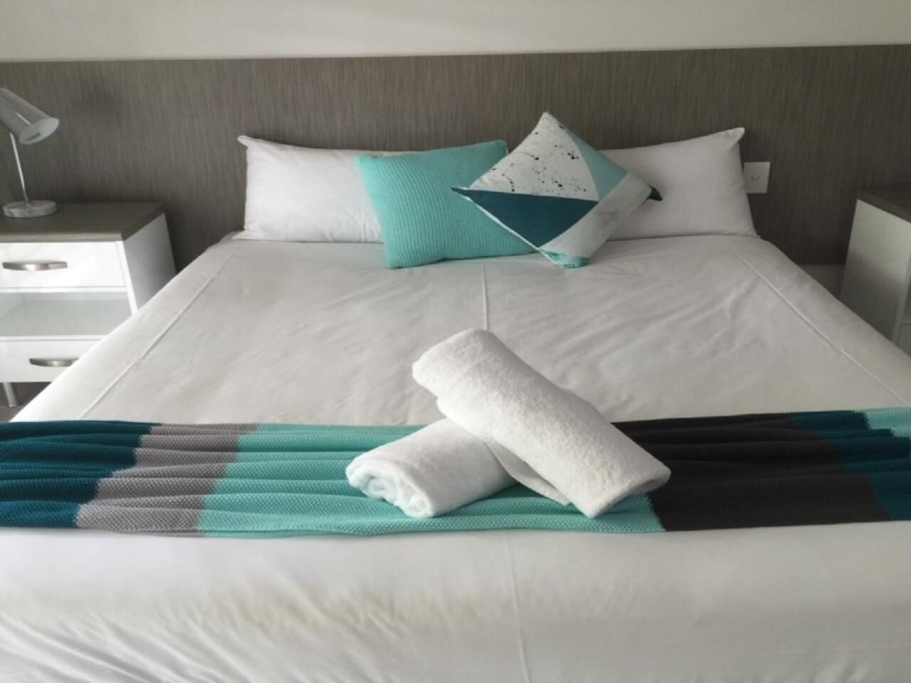 A neatly made bed with a white duvet, two pillows, a teal throw blanket, and two rolled towels.