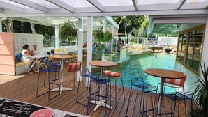 Un espace piscine extérieur doté de sièges et de tables, entouré de murs en verre et d’une terrasse en bois.