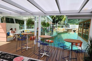 Un área de piscina al aire libre con asientos y mesas, rodeada por paredes de vidrio y una terraza de madera.