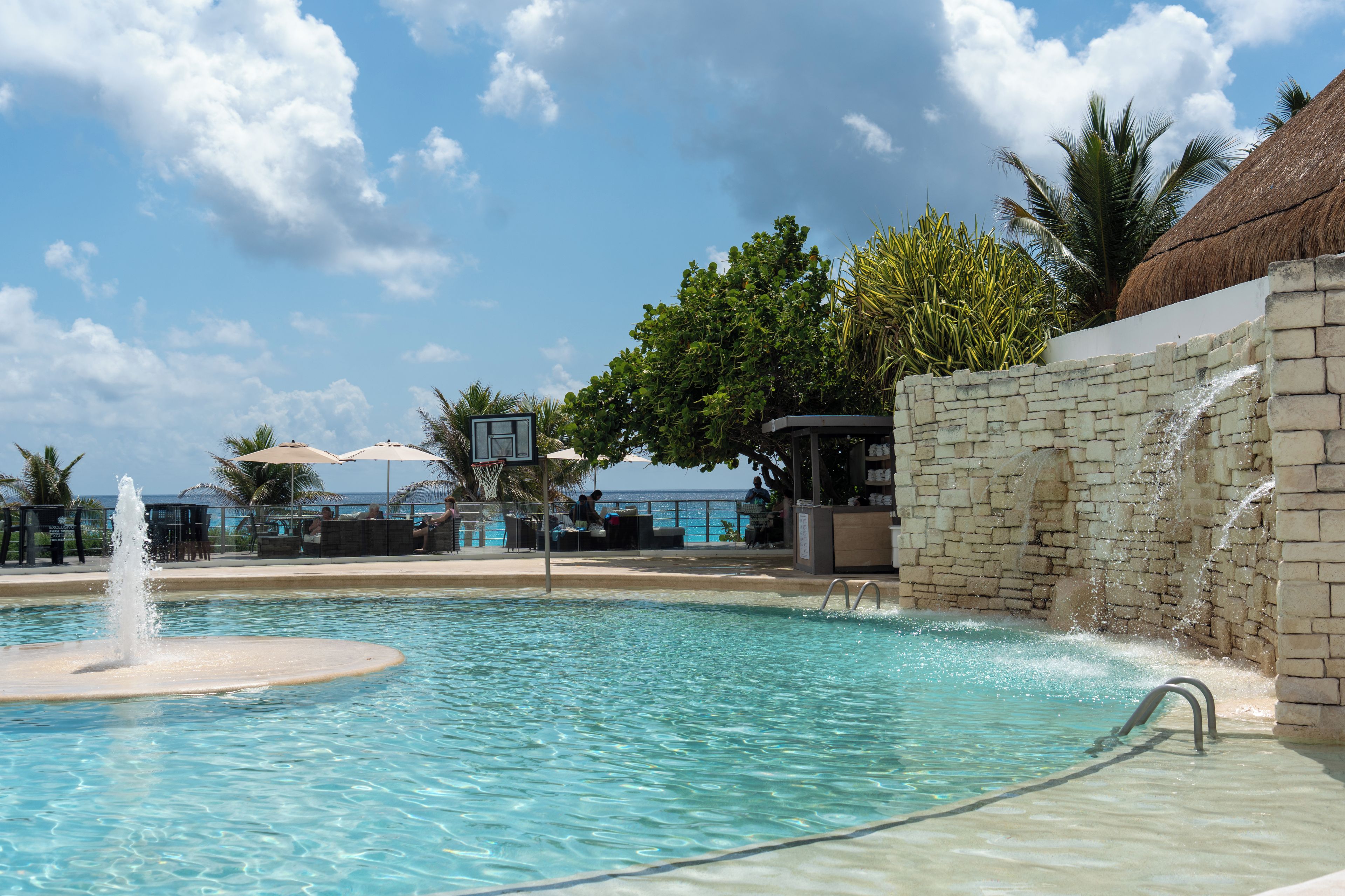 A clear pool with a water fountain, surrounded by lounge chairs and umbrellas, with a view of the ocean in the background.