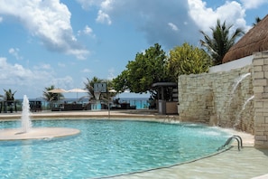 A clear pool with a water fountain, surrounded by lounge chairs and umbrellas, with a view of the ocean in the background. - Sunset Royal Beach Resort (Cancun)