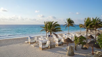 Una playa con sillas de playa blancas, palmeras y sombrillas de paja.