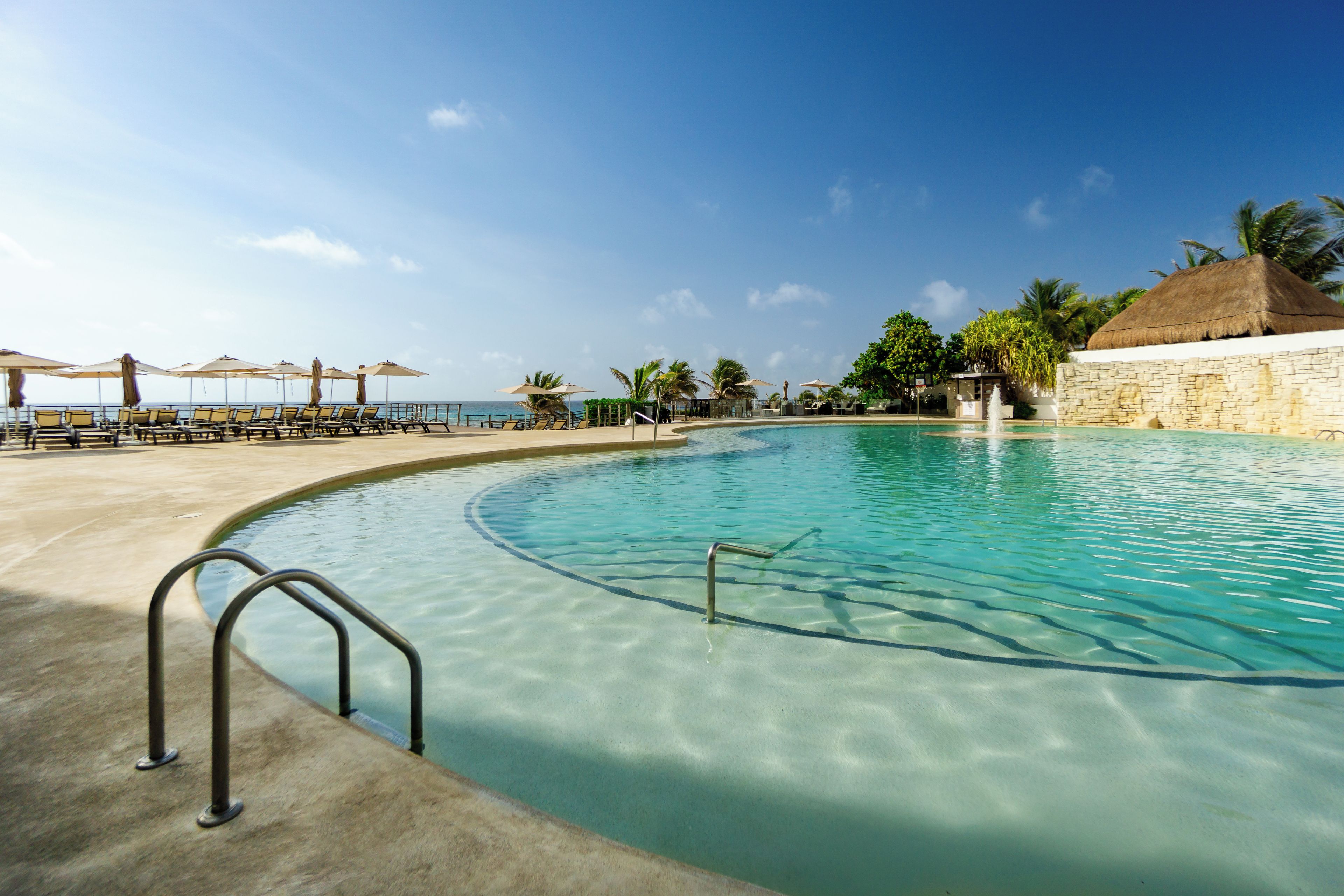 A clear blue pool with a curved edge, surrounded by lounge chairs and umbrellas, with a view of the ocean and palm trees in the background.