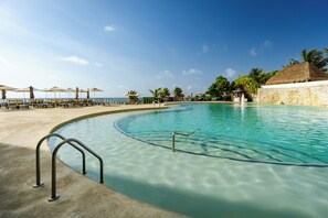 Una piscina azul cristalina con borde curvo, rodeada de sillas de descanso y sombrillas, con vistas al océano y palmeras al fondo.