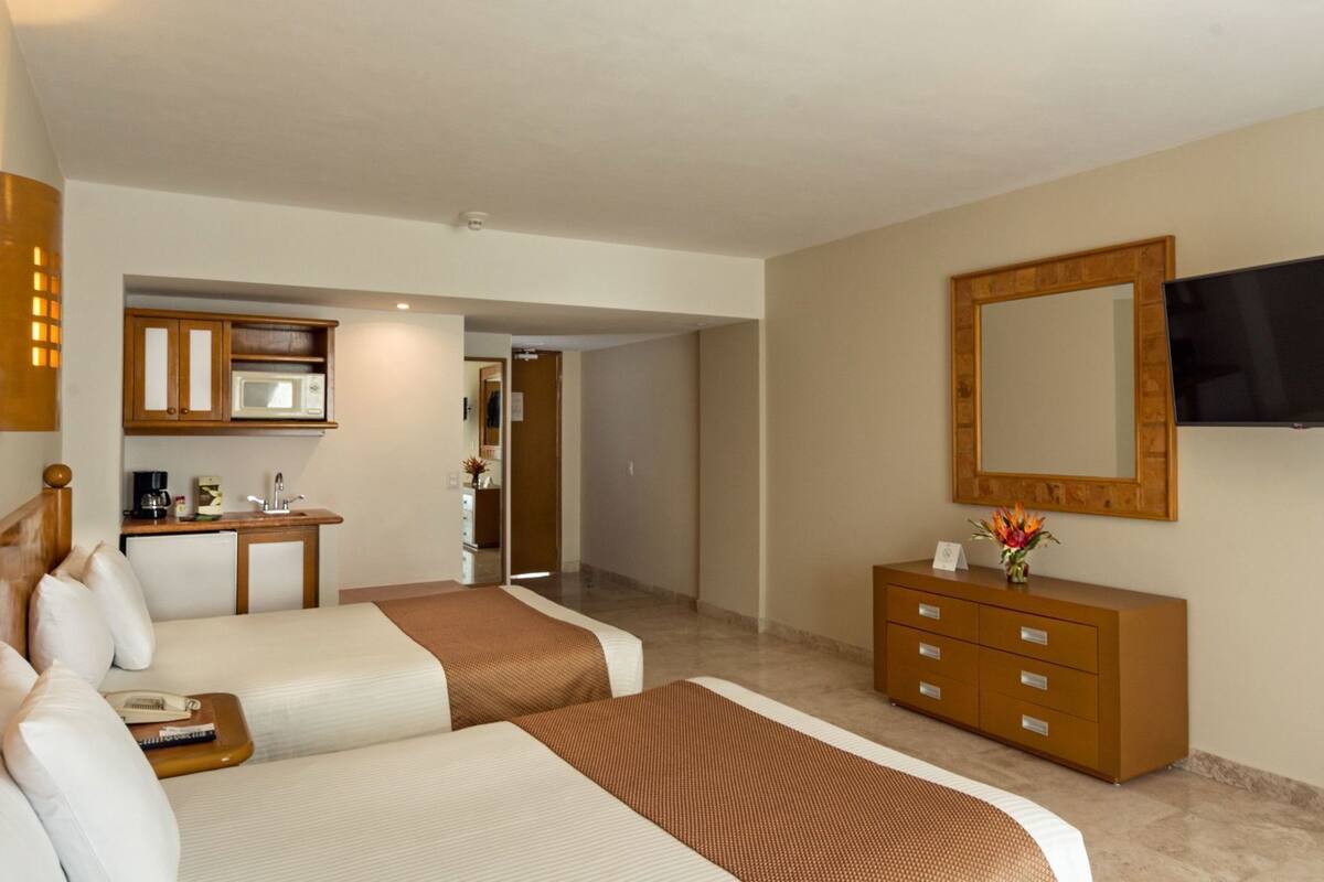 A hotel room with two beds, a small kitchenette, a bathroom, and a TV mounted on the wall.