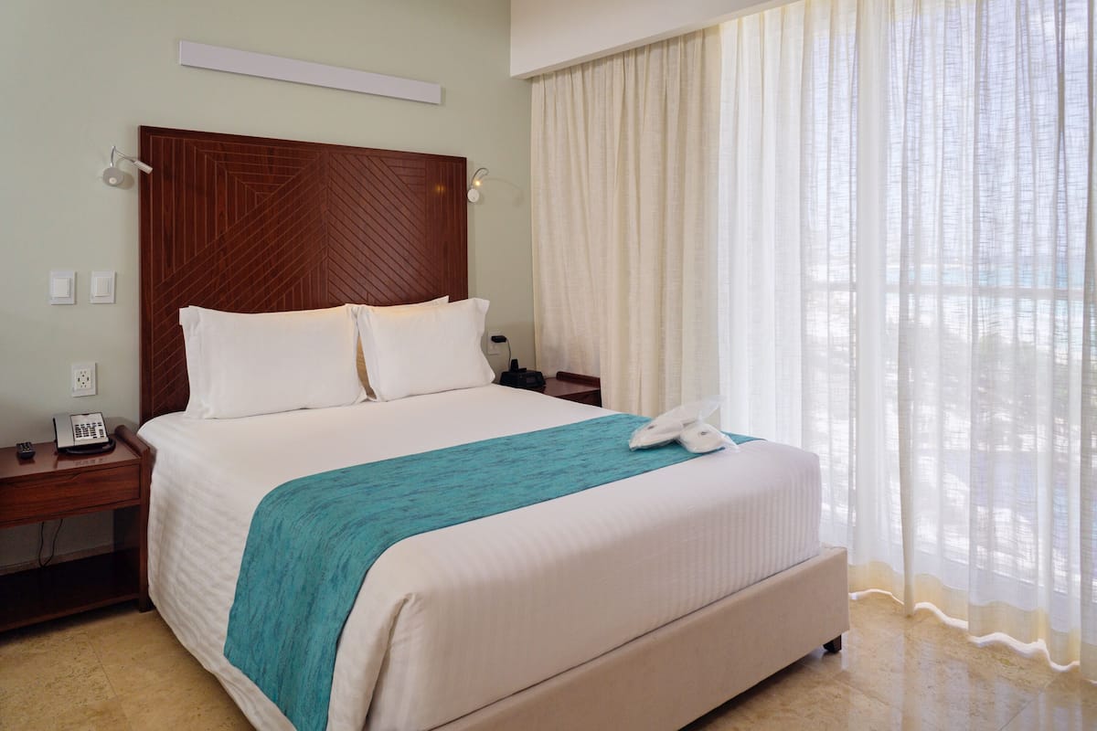 A hotel room with a large bed, a wooden headboard, a bedside table with a phone, and a window with sheer curtains.