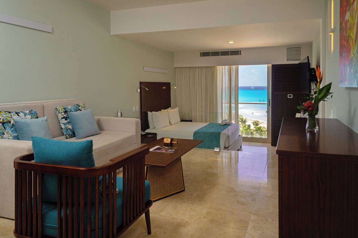 A hotel room with a balcony, a bed, a sofa, a wooden chair, and a desk.
