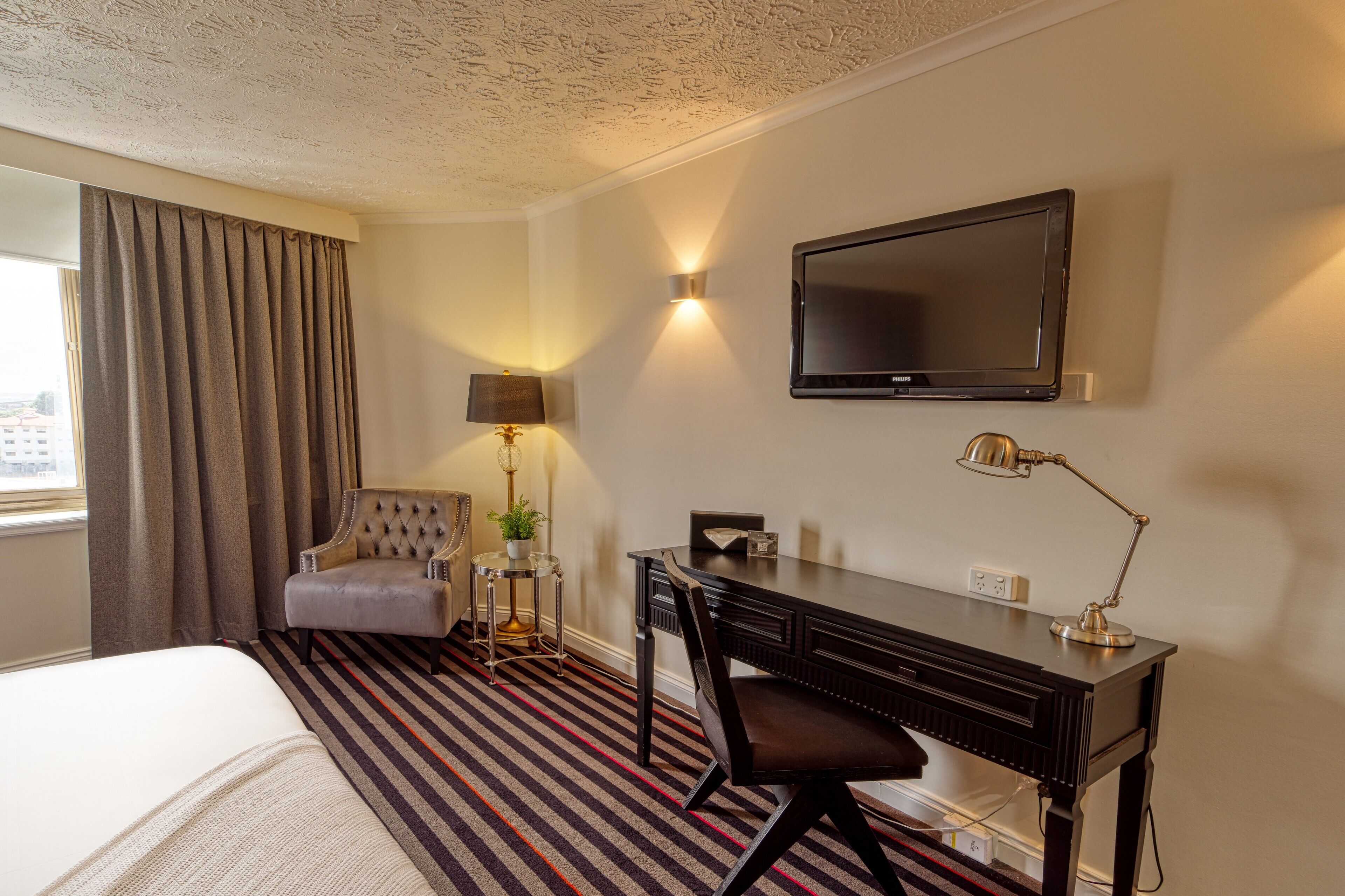 A hotel room with a bed, a desk, a chair, a TV, and a lamp.