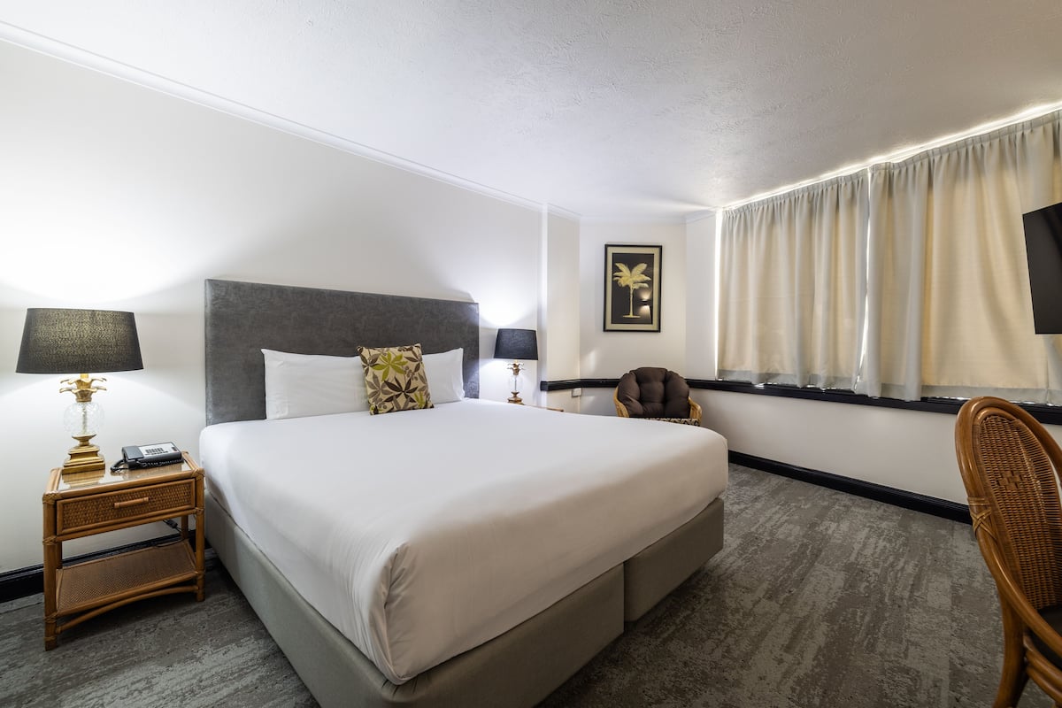 A hotel room with a large bed, two bedside tables with lamps, a chair, a window with curtains, and a wall-mounted TV.