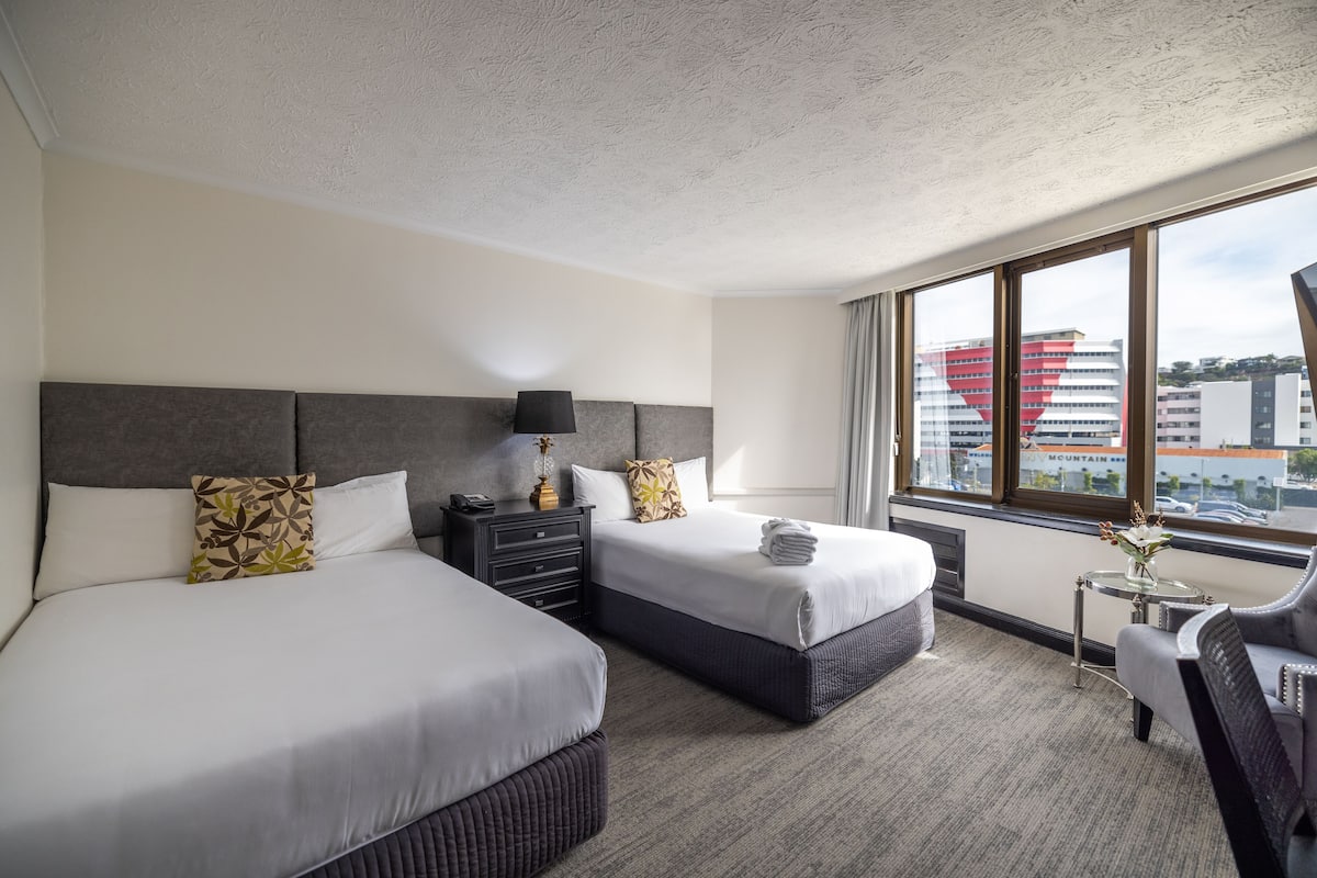 A hotel room with two beds, a nightstand, a lamp, a chair, and a view of buildings through large windows.