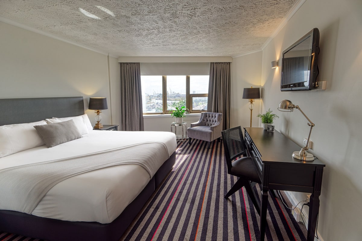 A hotel room with a large bed, a desk with a chair, a television, and a view of the city through the window.