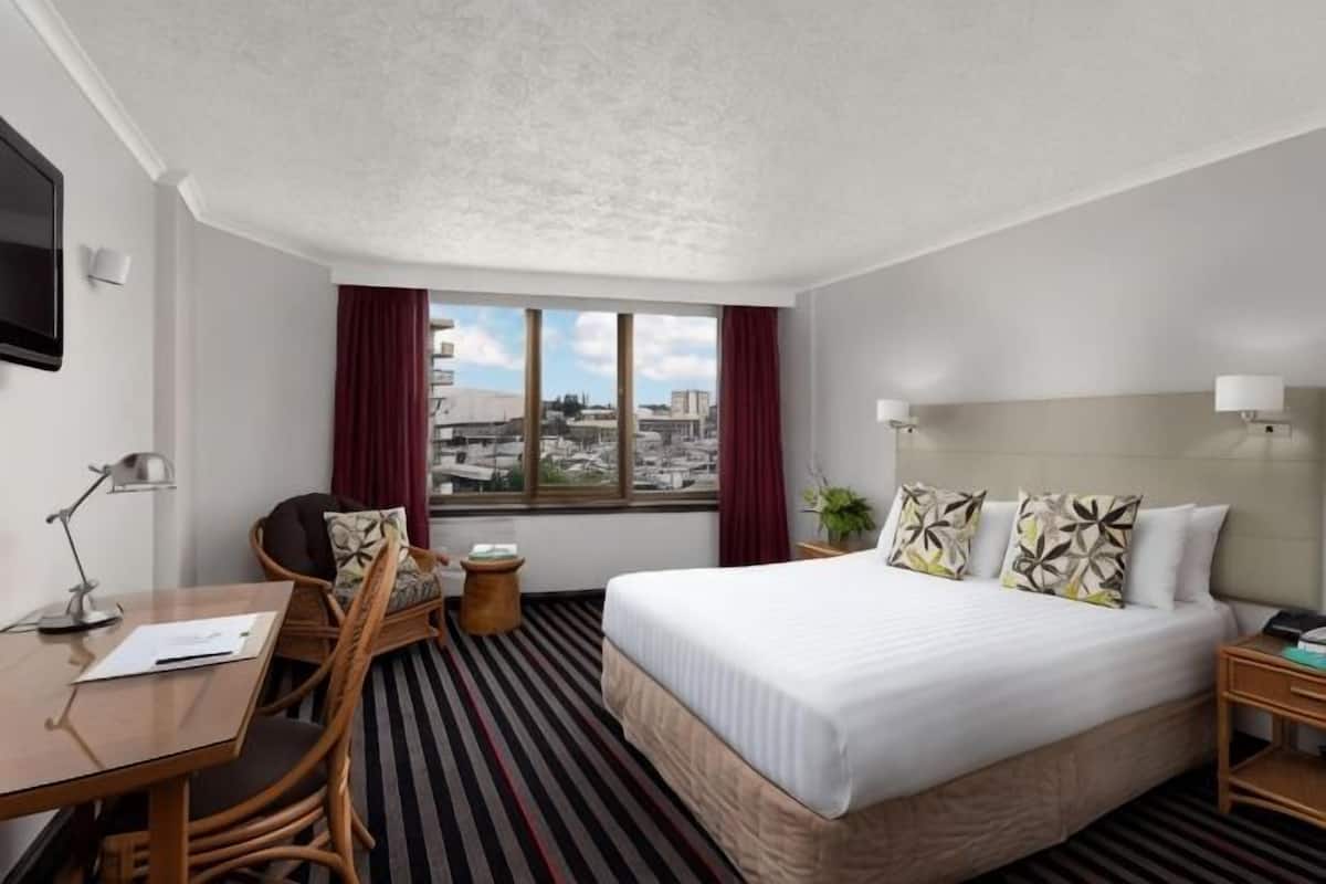 A hotel room with a bed, a desk with a chair, a small table, a TV, and a window with curtains.