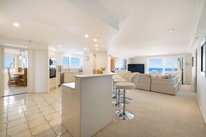 Una sala de estar moderna con un sofá, un comedor con sillas, una cocina con lavaplatos y estufa, y ventanas grandes con vistas.