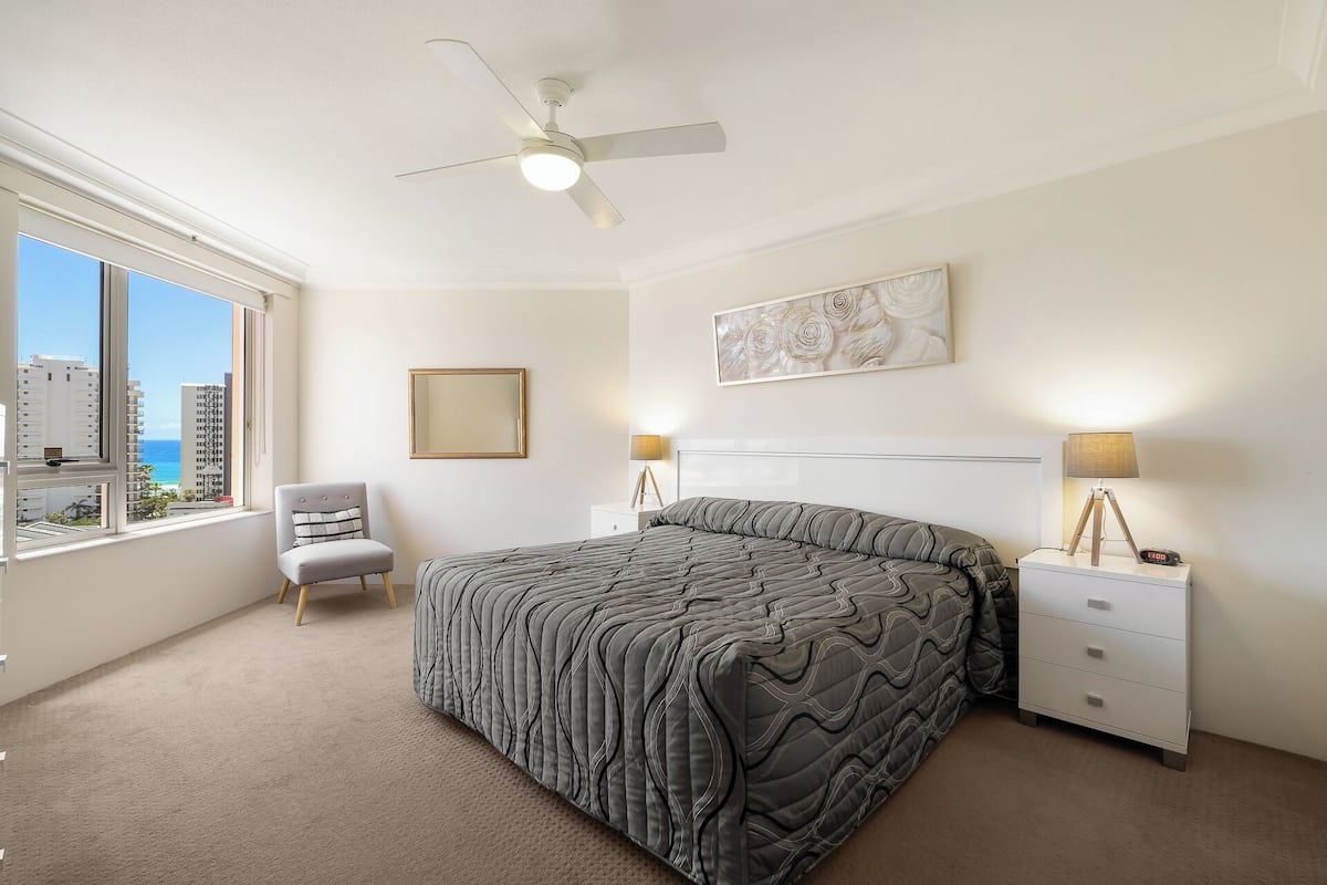 A bedroom with a bed, a chair, a nightstand, a lamp, a ceiling fan, a window with a view of buildings, and a framed picture on the wall.
