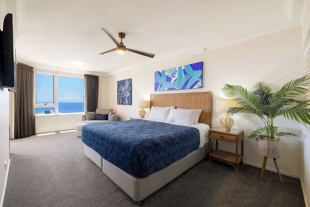 A hotel room with a bed, a ceiling fan, a large window with a view, a painting, and a potted plant.