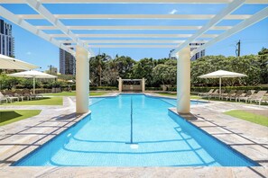 Una piscina rectangular con borde de azulejos, rodeada de sillas de descanso y sombrillas, bajo una estructura de pérgola blanca.