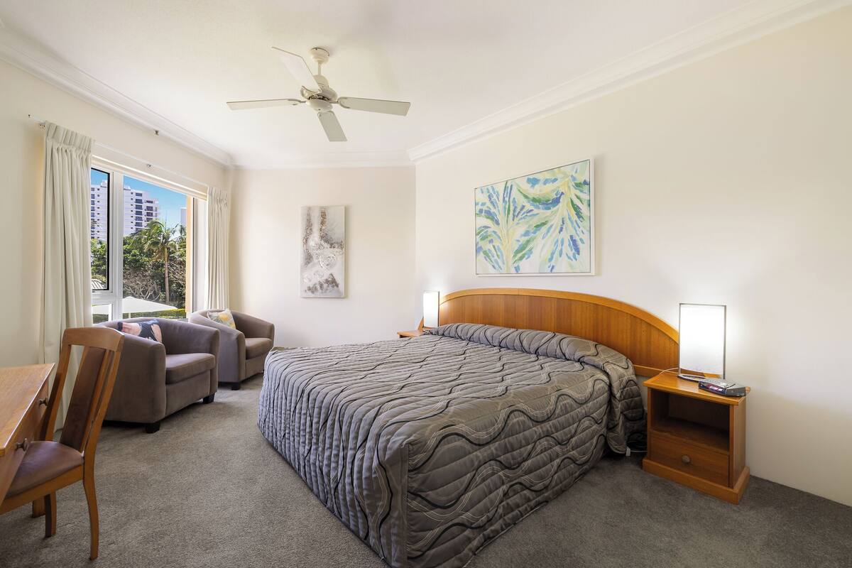 A hotel room with a bed, a chair, a desk, a nightstand, a ceiling fan, and artwork on the walls.