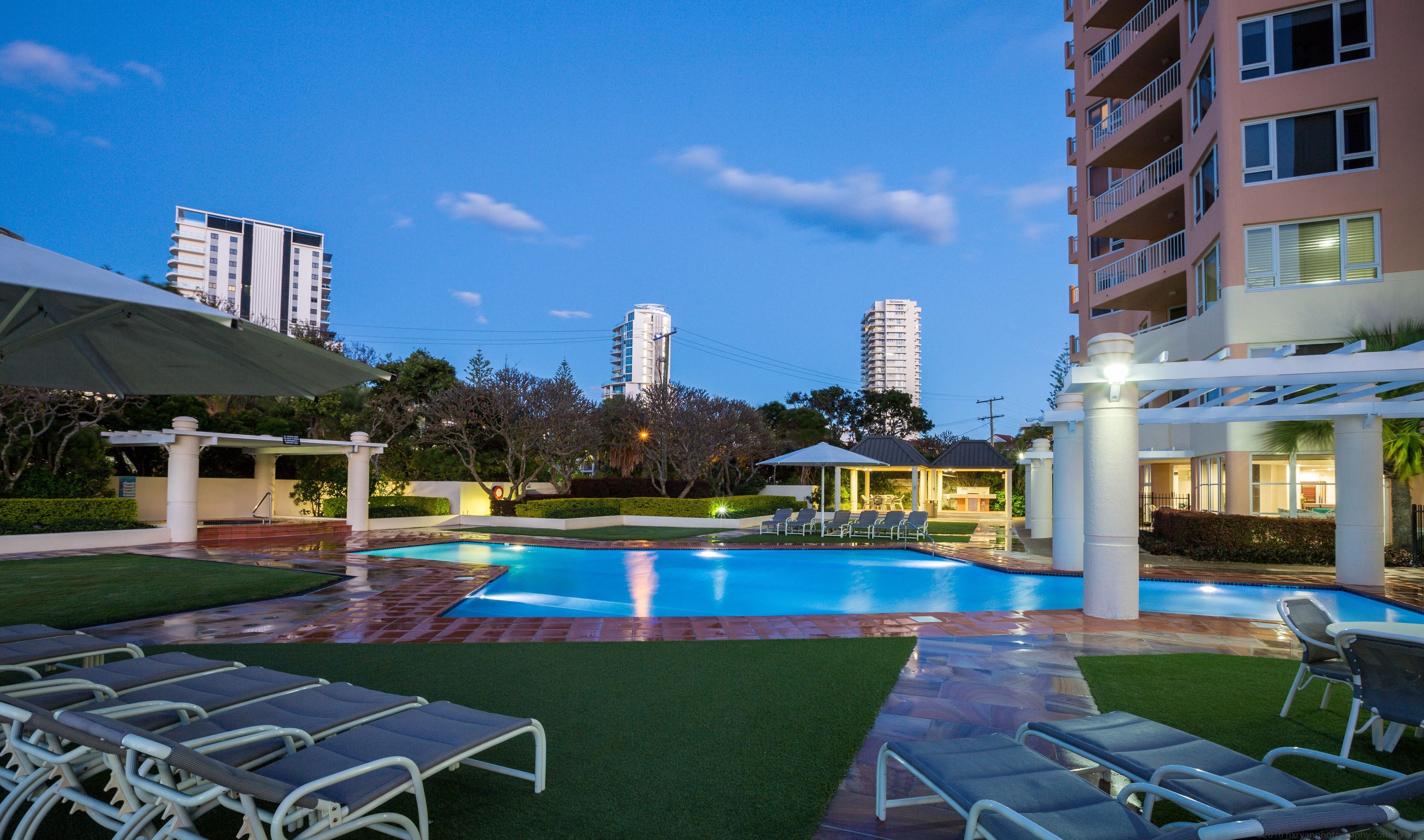 A swimming pool with lounge chairs, a covered seating area, and a view of tall buildings in the background.