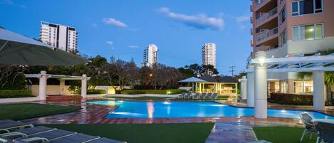 A swimming pool with lounge chairs, a covered seating area, and a view of tall buildings in the background.