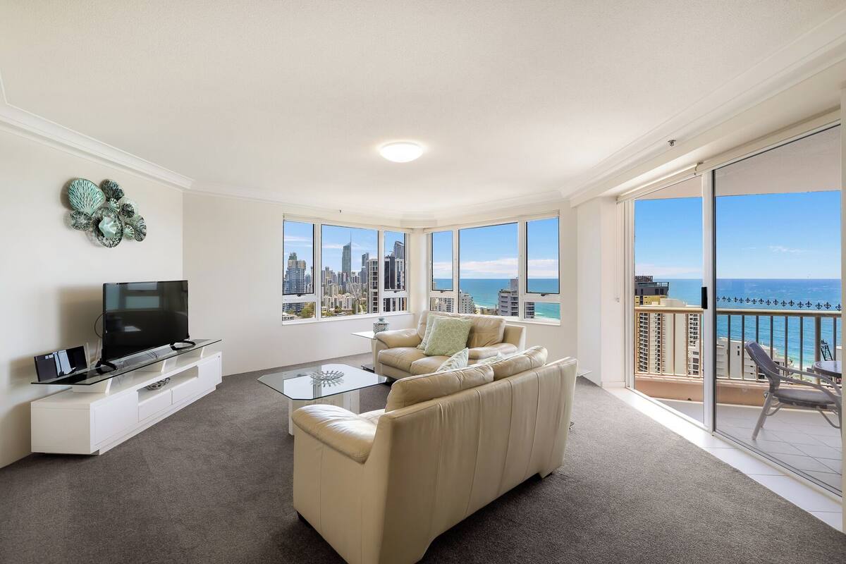 A modern living room with a sofa, TV, and glass coffee table. Large windows offer a cityscape and ocean view.