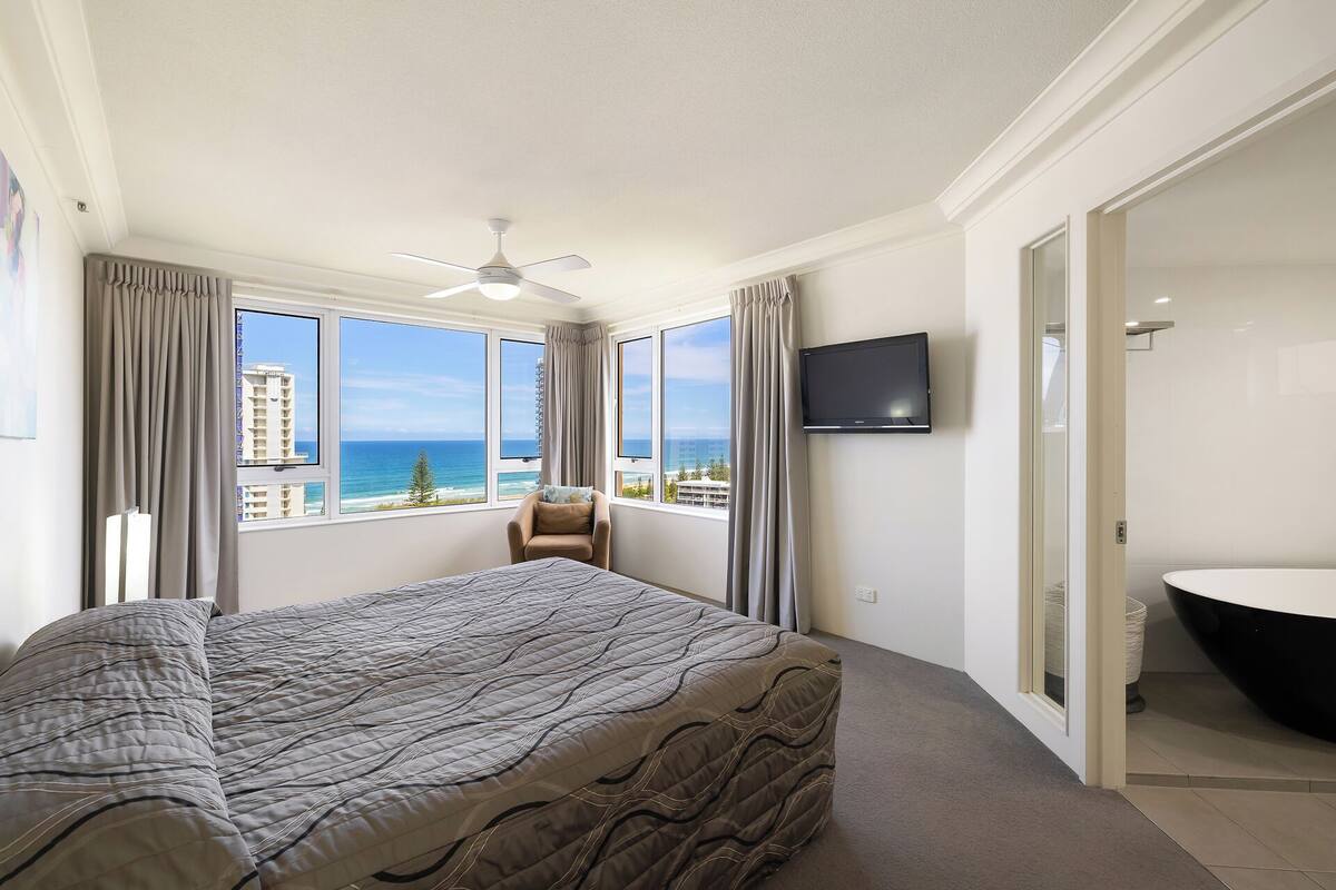 A hotel room with a bed, a TV, a bathroom, and a view of the ocean.