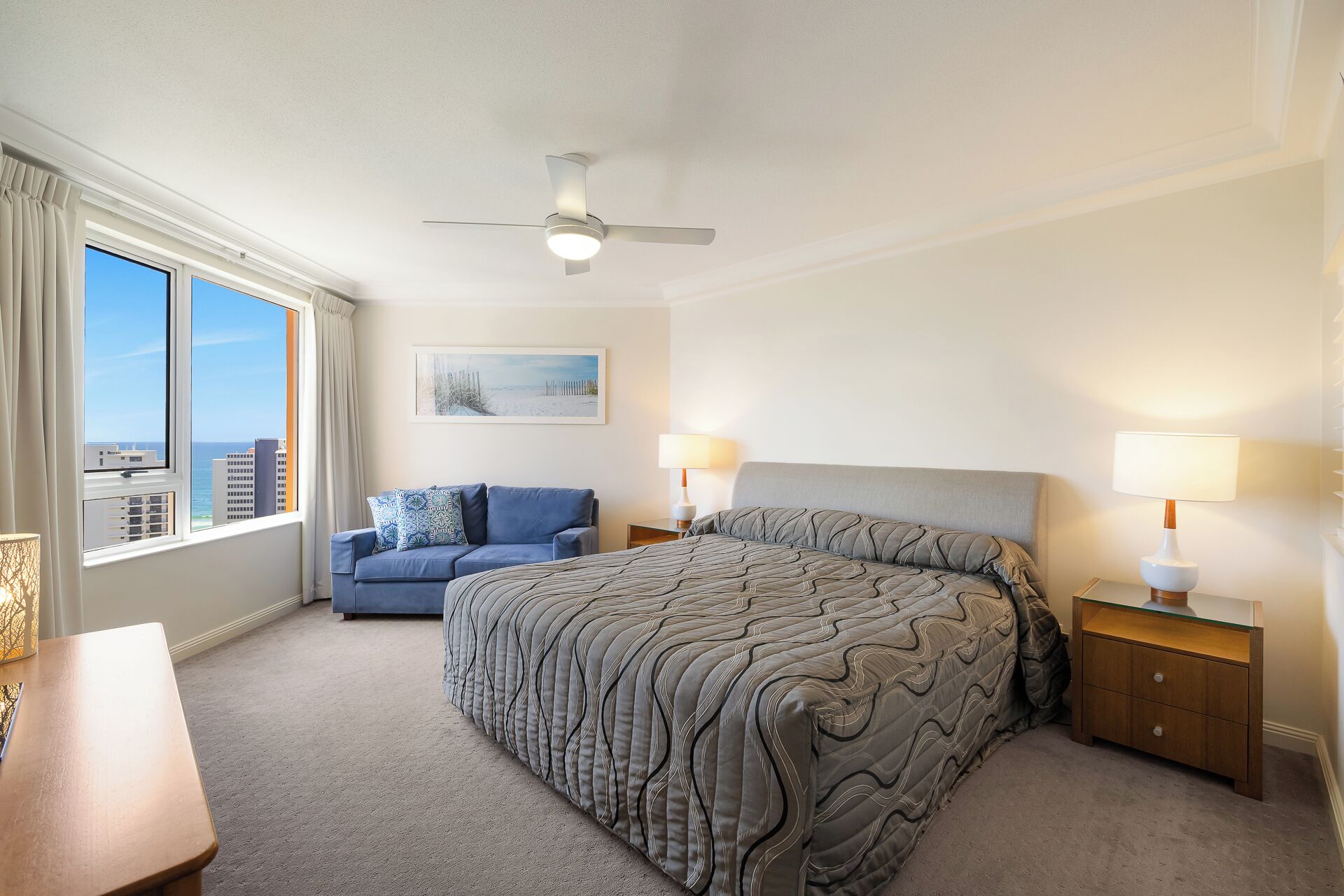 A bedroom with a bed, a nightstand, a lamp, a sofa, a ceiling fan, and a window with a view of buildings and the sky.