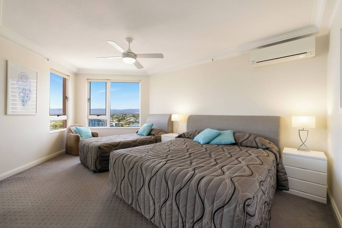 A bedroom with two beds, a ceiling fan, an air conditioning unit, and a window with a city view.