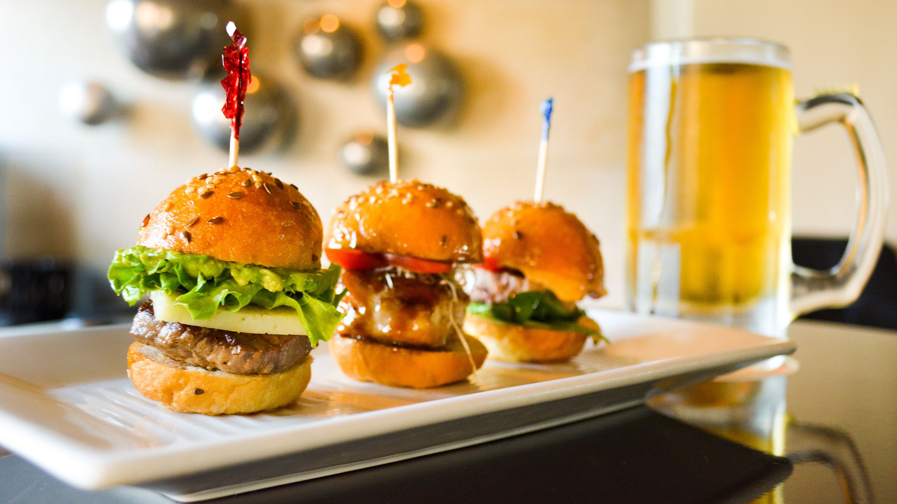 Three burgers with toothpicks on a white plate, and a glass mug with a yellow beverage in the background.