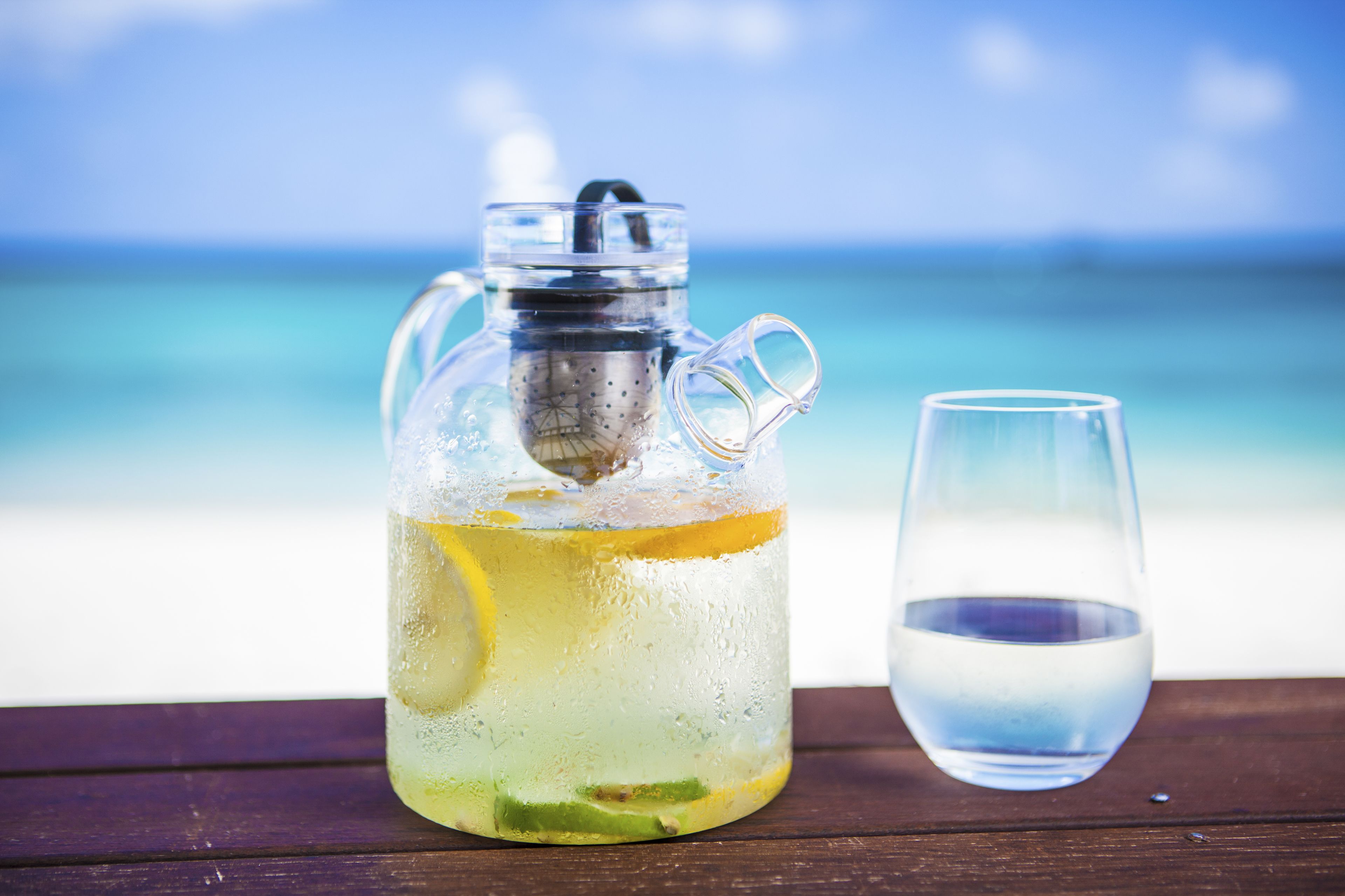A glass pitcher with a handle and a glass beside it on a wooden surface with a beach and ocean in the background.