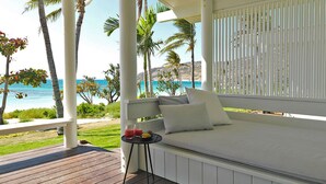 Un balcón con zona de estar acolchada de color blanco, una pequeña mesa con una bebida y vista al océano y palmeras.