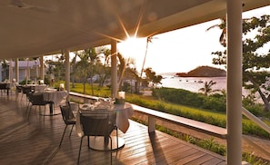 An outdoor dining area with tables and chairs, overlooking a coastal view with palm trees and boats.