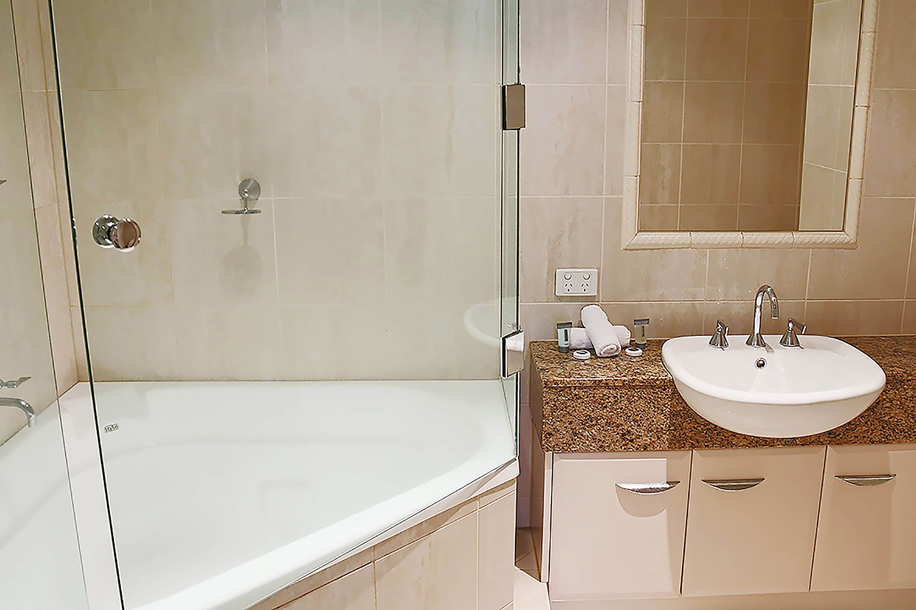 A bathroom with a glass-enclosed shower, a white bathtub, a sink with a granite countertop, and beige cabinetry.