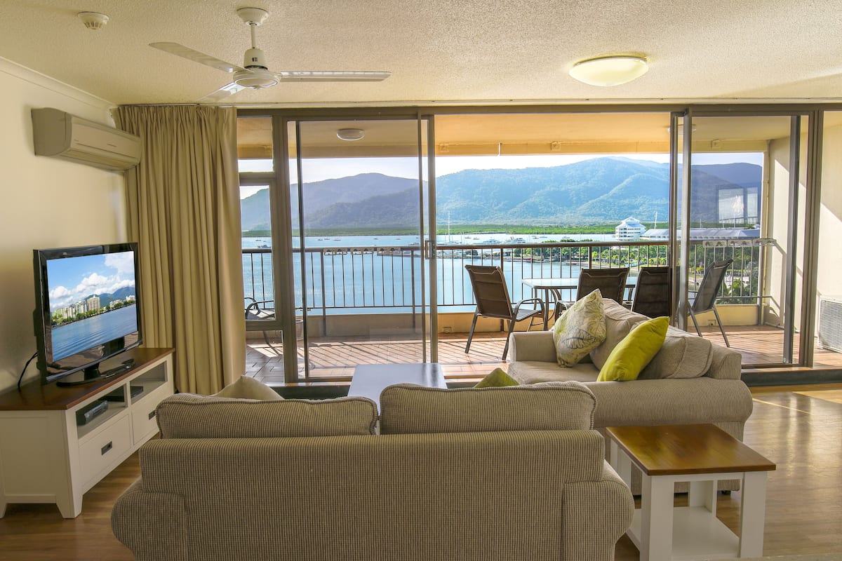 Una sala de estar con televisor, sofá y un balcón con vistas al mar y a las montañas.