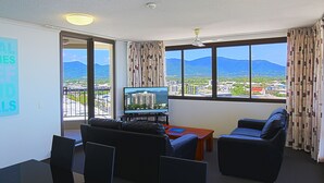 Una sala de estar con sofá, sillas, una mesa de centro, televisor y vistas a montañas a través de amplios ventanales.