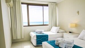 Habitación de hotel con dos camas, una ventana que da al mar, una mesita de noche con lámpara y toallas sobre las camas.