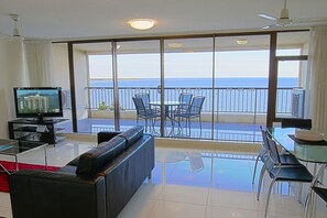 Una sala de estar con una televisión grande, un sofá, una mesa de centro de vidrio y un balcón con una mesa y sillas.