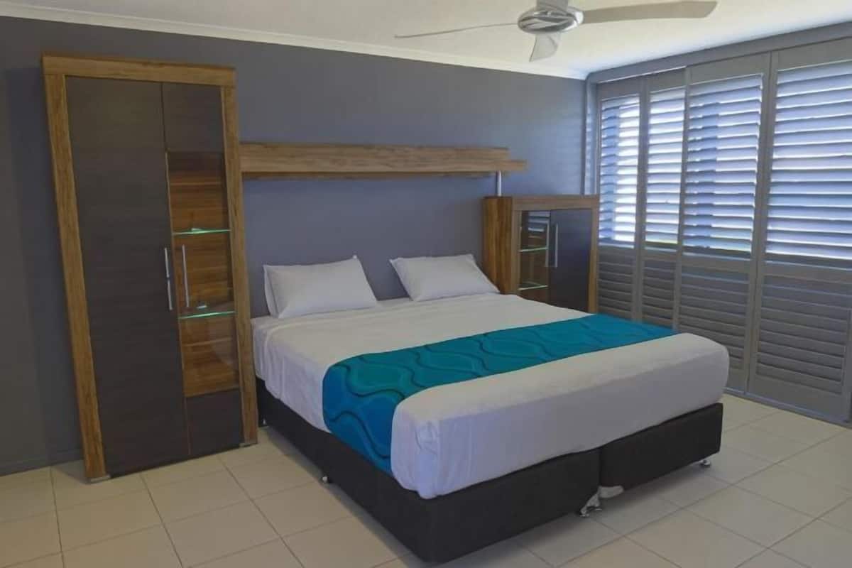 Un dormitorio con cama, armario, mesitas de noche y una ventana con persianas.