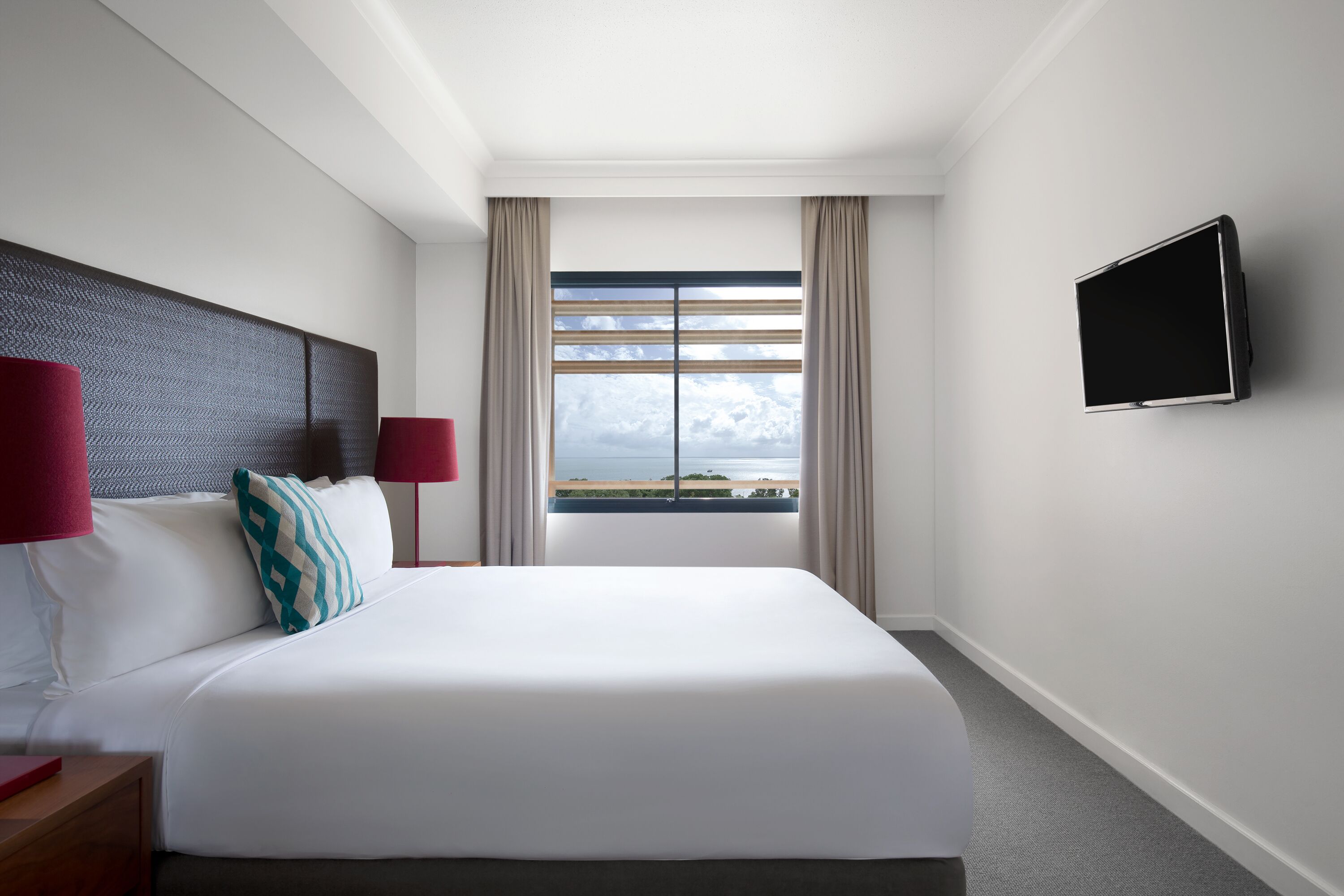 A hotel room with a large bed, a flat-screen TV mounted on the wall, and a window with a view of the sea.