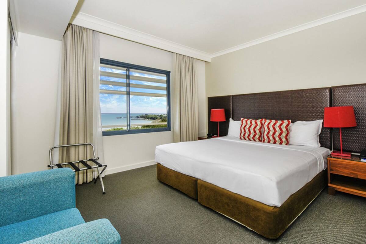 A hotel room with a large bed, two bedside tables with lamps, a chair, a window with curtains, and a view of the sea.