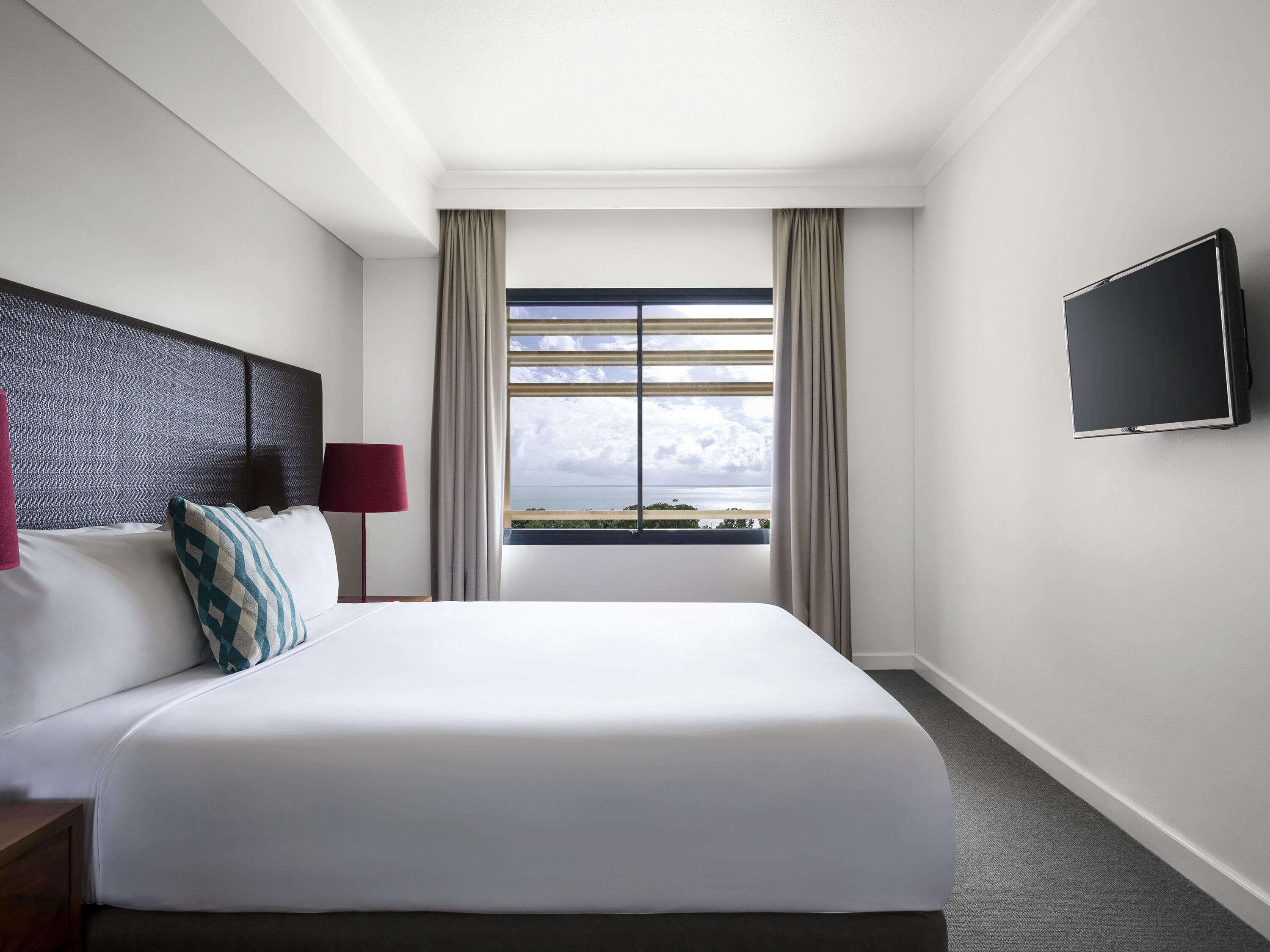 A hotel room with a large bed, a flat-screen TV, a window with curtains, and a view of the sea.