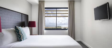 A hotel room with a large bed, a flat-screen TV, a window with curtains, and a view of the sea.
