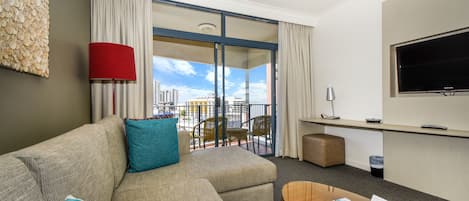 Habitación de hotel con sofá, mesa de centro, televisor, balcón con vistas y escritorio.